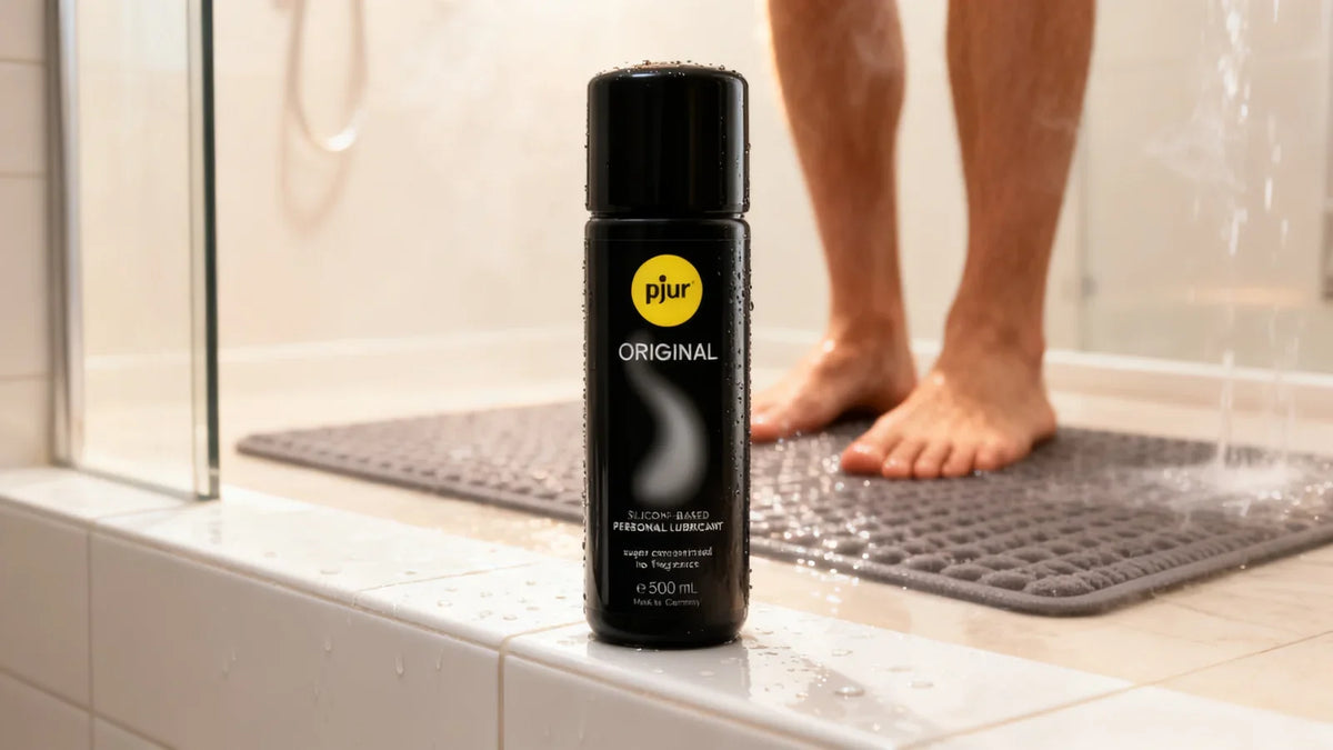 Flacon noir de lubrifiant silicone Pjur Original posé sur le rebord carrelé d’une douche à l’italienne, gouttes d’eau et vapeur, tapis antidérapant et pieds nus flous à l’arrière-plan